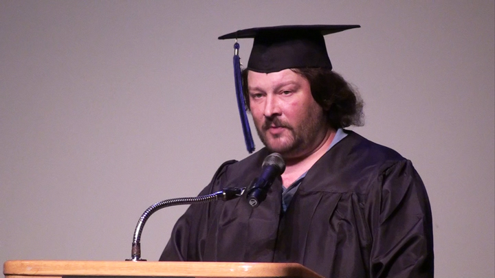 Man in graduation cap and gown speaking into microphone at podium