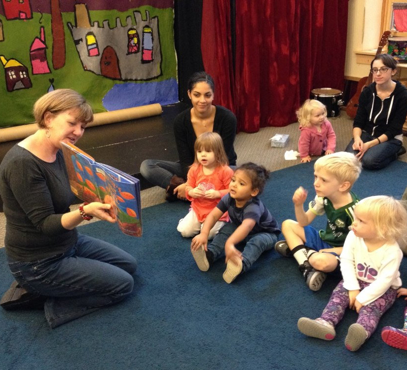 Children sitting on floor in front of adult reading them a book