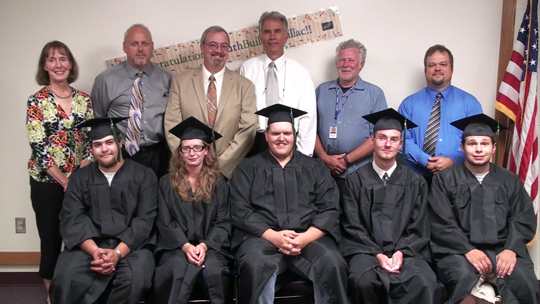 Two rows of people. In the back they are older and standing. In the front are young a wearing graduation caps and gowns